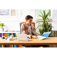 Ein junger Mann sitzt an einem Schreibtisch, arbeitet mit einem Laptop und blättert durch Unterlagen, umgeben von Pflanzen und bunten Ordnern.