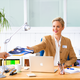 Eine Frau in einem Büro überreicht einem Mann ein Dokument, während sie an einem Schreibtisch mit einem Laptop und Büromaterial arbeitet.