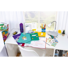 Ein Schreibtisch mit bunten Ordnern, Schreibmaterialien, einer Lampe und einer Blume. Im Hintergrund sind Fenster und ein farbenfrohes Aufbewahrungselement sichtbar.