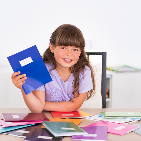 Ein Mädchen hält ein blaues Heft in der Hand, umgeben von mehreren bunten Heften auf einem Tisch.