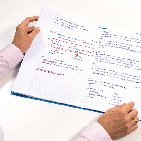 Eine Person hält ein aufgeschlagenes Notizbuch mit handschriftlichen Notizen und Markierungen in blauer und roter Tinte." 

(Note: The original text is already in German and does not require translation.)