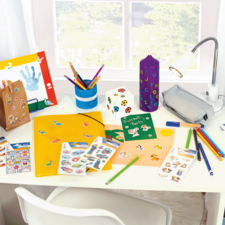 Bunt geschmückter Schreibtisch mit Bastelmaterialien, Aufklebern, Stiften, einem Ordner und einer Lampe.