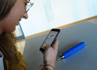 Eine Person schaut auf ihr Smartphone, während ein blauer Roller auf dem Tisch steht.