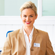 Eine lächelnde Frau mit blonden Haaren trägt einen beigen Blazer und eine weiße Bluse. Ihr Namensschild mit der Aufschrift 'Manuela Richter' ist gut zu erkennen.