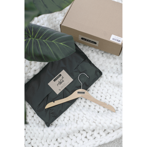 Ein schwarzes Paket mit einem Etikett und einem Holzkleiderbügel liegt auf einer weißen Decke, umgeben von einer Pflanze.