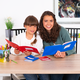Zwei Mädchen sitzen an einem Tisch und lesen Bücher. Eines hat ein rotes Buch, das andere ein blaues. Buntstifte und Notizblätter liegen auf dem Tisch.