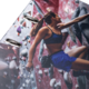 Eine Kletterin in Sportbekleidung erklimmt eine Kletterwand mit bunten Griffen. Sie konzentriert sich auf ihren nächsten Halt.