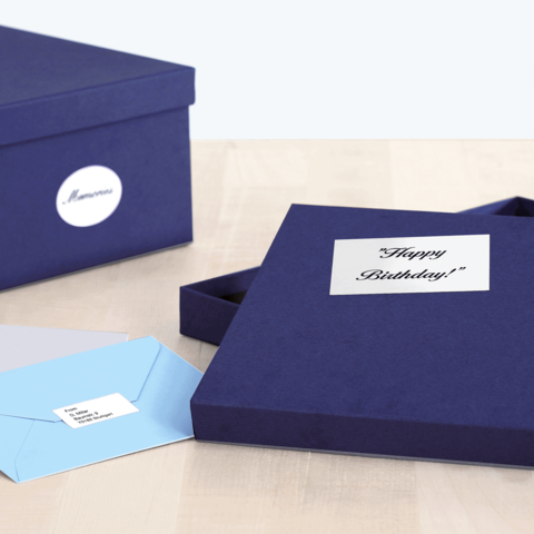 Eine geöffnete, dunkelblaue Geschenkbox mit der Aufschrift "Happy Birthday!" und zwei Umschlägen in hellblau und grau daneben.