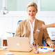 Eine Frau mit blonden Haaren sitzt an einem Schreibtisch, trägt einen Blazer und lächelt. Vor ihr steht ein Laptop und Schreibutensilien.