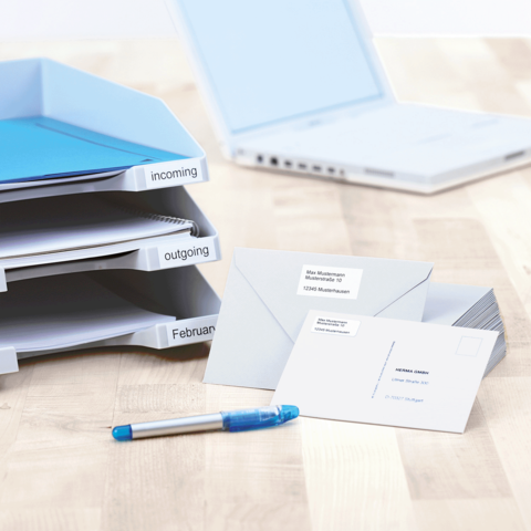 Ordner mit beschrifteten Fächern für eingehende und ausgehende Dokumente, daneben ein Laptop, Umschläge und ein blauer Stift.