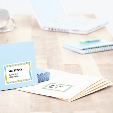 Blaue und weiße Briefumschläge mit Adressaufklebern liegen auf einem Tisch, neben einem Laptop und einem Notizbuch.