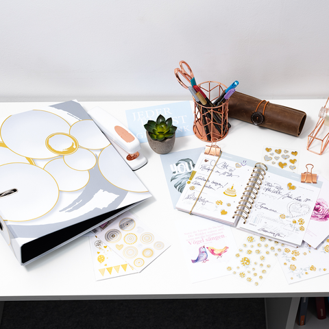 Schreibtisch mit Planer, Schreibutensilien, einer Pflanze und verschiedenen Dekorationselementen. Farbiges Papier und Aufkleber sind verteilt.