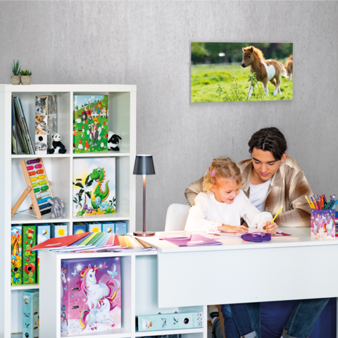 Ein Mädchen sitzt an einem Schreibtisch, malt mit Stiften und hat eine Federmappe vor sich. Im Hintergrund sind Regale mit Spielzeug und Kunstmaterialien.