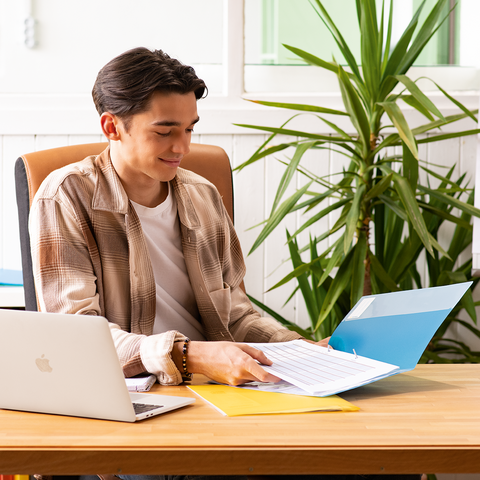 Ein junger Mann sitzt an einem Schreibtisch, arbeitet mit einem Laptop und blättert durch Dokumente in einer Ordner. Pflanzen und farbige Aktenordner sind im Hintergrund.