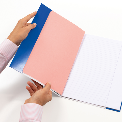 Eine Person hält ein geöffnetes Notizbuch mit einem blauen und rosa Einband, dessen leere linierten Seiten sichtbar sind.