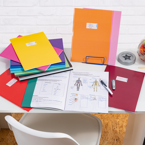 Bunte Ordner und Dokumente auf einem Schreibtisch, daneben ein Glas mit Süßigkeiten und Bürobedarf wie einen Hefter und einen Stift.