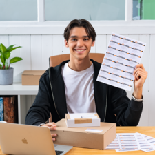 Ein junger Mann sitzt an einem Tisch, lächelt und hält ein Blatt mit Aufklebern in der Hand. Vor ihm stehen ein Karton und ein Laptop.