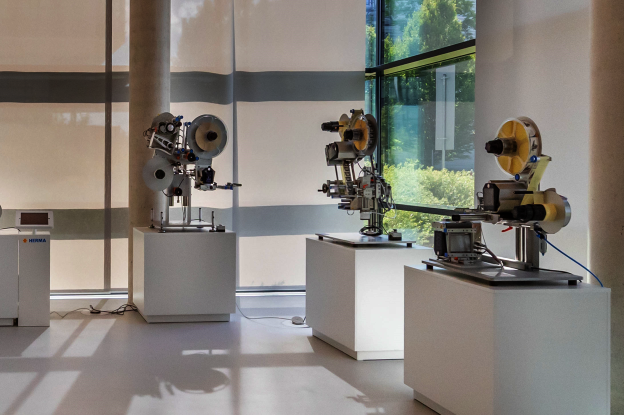 Ausstellung von Maschinenmodellen auf weißen Podesten in einem modernen Raum mit großen Fenstern und natürlichem Licht.