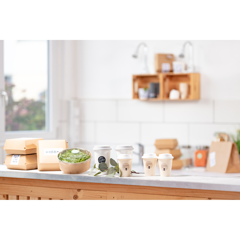 Eine Holzschüssel mit Salat, mehrere Kaffeebecher und Takeaway-Boxen auf einer Küchentheke in einem hellen Raum.