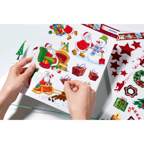 Hände halten ein Blatt mit bunten Weihnachtsstickern, darunter Weihnachtsmänner, Geschenke, ein Schneemann und Weihnachtsdekoration.