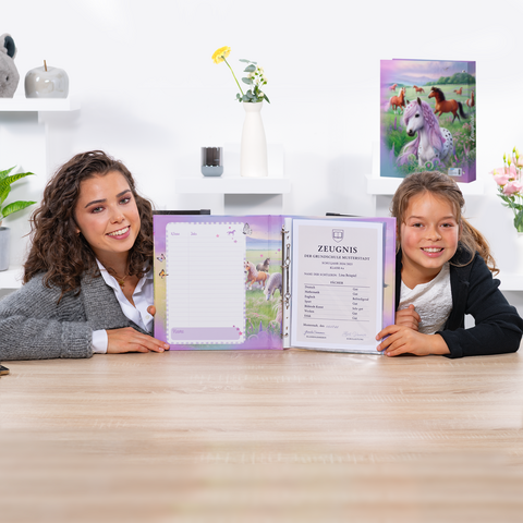 Zwei Mädchen sitzen an einem Tisch und halten ein offenes Heft mit einem Zertifikat und einem leeren Blatt. Im Hintergrund sind dekorative Elemente zu sehen.