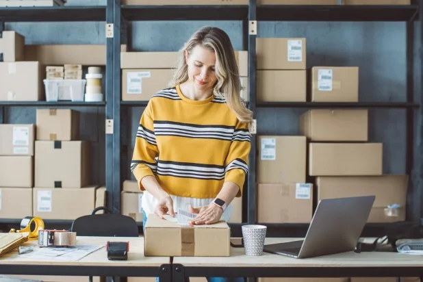 Eine Frau in einem gestreiften Pullover steht an einem Tisch, umgeben von Kartons und einem Laptop in einem modernen Büro."
(Note: The text is already in German and does not require translation. It is clear and fluent as it is.)