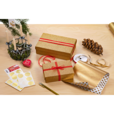 Geschenkpäckchen in goldener Verpackung mit roten Bändern, umgeben von Geschenkpapier, Aufklebern, einer Tannenzapfen und Kerzenhalter.