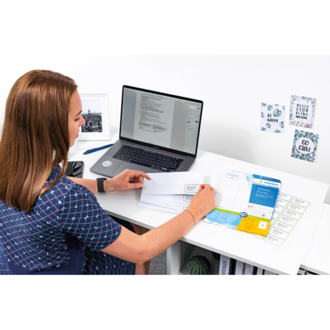 Eine Person sitzt an einem Schreibtisch, arbeitet am Laptop und sortiert Dokumente sowie Briefe.