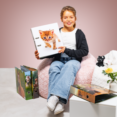 Ein Mädchen sitzt auf einem Sitzsack und hält einen Ordner mit einem Kätzchen auf dem Cover. Rund um sie stehen weitere Ordner und eine Pflanze.