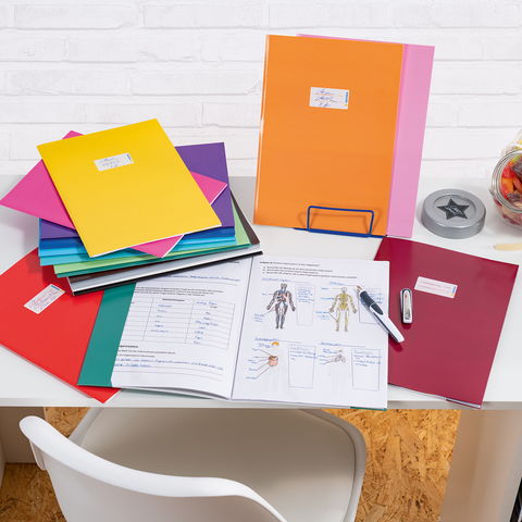 Bunte Ordner und Unterlagen auf einem Schreibtisch, daneben ein Glas mit Süßigkeiten und Bürobedarf wie ein Hefter und ein Stift.