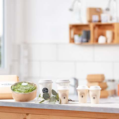 Ein Holztisch mit einer Schüssel Salat, mehreren Kaffeetassen und einer Verpackung, umgeben von einer hellen, modernen Küche.