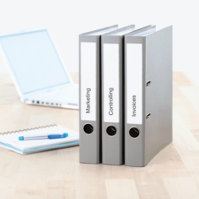 Drei graue Ordner mit den Beschriftungen "Marketing", "Controlling" und "Rechnungen" stehen auf einem Tisch neben einem Laptop und einem Notizblock.