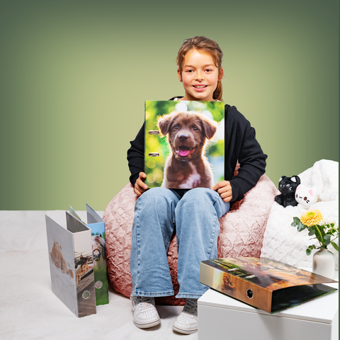 Ein Mädchen sitzt auf einem Pouf und hält einen Ordner mit einem Hundebild. Neben ihr liegen weitere Ordner und eine kleine Pflanze." 

(Note: The text is already in German and does not require translation. It is clear and fluent as it is.)