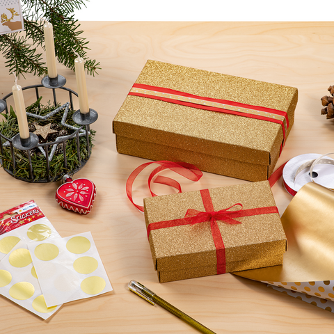 Geschenkverpackungen in Gold und Weiß mit Punkten, umgeben von Dekorationen wie Kerzen, Tannenzapfen und Geschenkband auf einem Holztisch.
