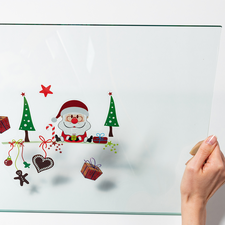 Eine transparente Glasplatte wird von zwei Händen gehalten, mit festlichen Motiven wie einem Weihnachtsmann, Tannenbäumen und Geschenken.