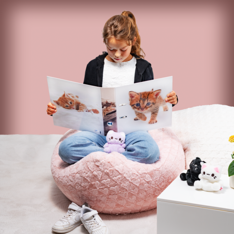 Ein Mädchen sitzt in einem großen, weichen Sitzsack und schaut sich ein Bilderbuch mit Kätzchen an. Um sie herum liegen Plüschtiere.