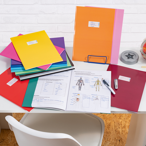 Bunte Ordner und Unterlagen liegen auf einem Schreibtisch, daneben ein Stift, ein Hefter und ein Glas mit Süßigkeiten.