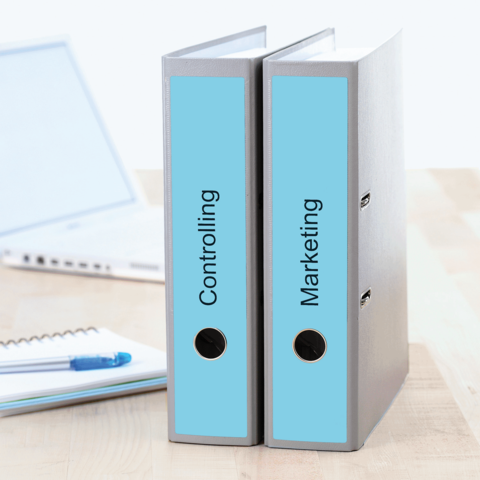 Zwei Ordner mit den Beschriftungen "Controlling" und "Marketing" stehen auf einem Tisch, neben einem Laptop und einem Notizblock.