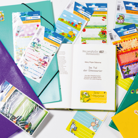 Hände halten einen bunten Notizzettel mit einem Cartoon-Motiv, umgeben von Schulmaterialien, einem Buch und einer Vase mit Blumen.