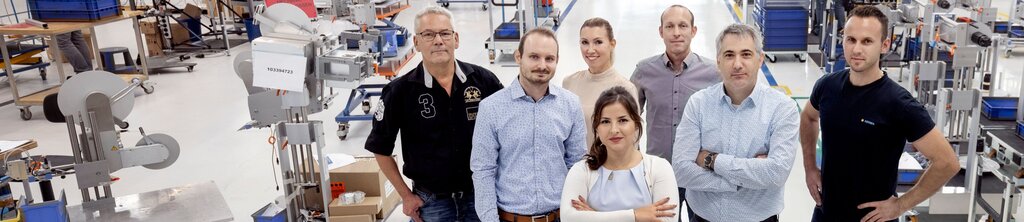 Eine Gruppe von sieben Personen steht in einer Fabrikhalle, umgeben von Maschinen und Arbeitsstationen. Sie blicken in die Kamera.