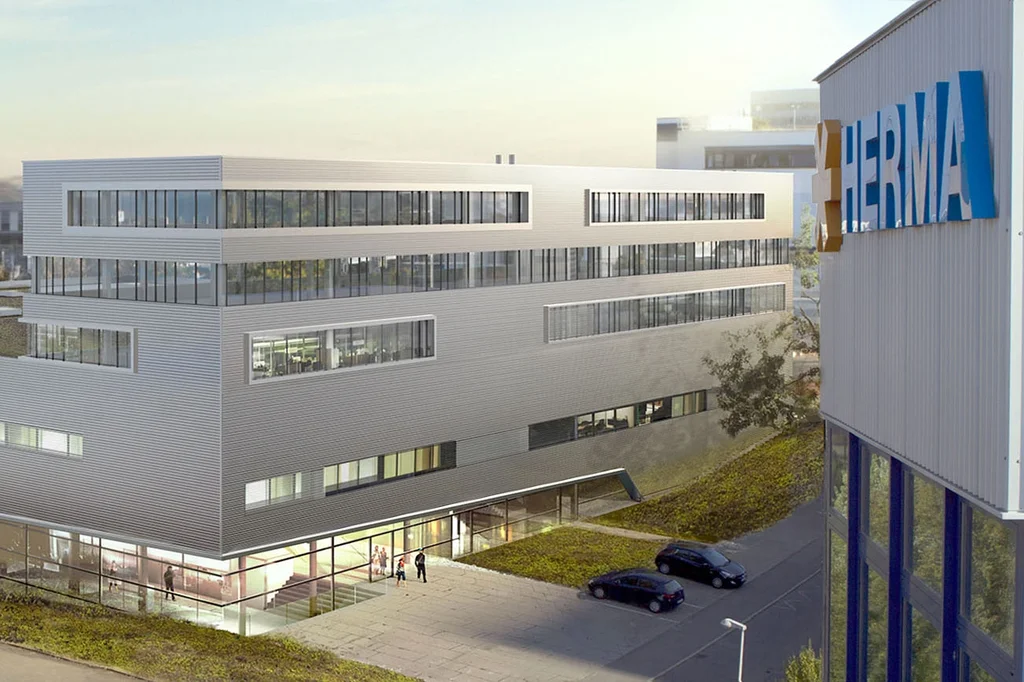 Moderne Bürogebäude mit flacher Dachlandschaft und großen Fenstern, umgeben von einer grünen Fläche und einem Parkplatz.