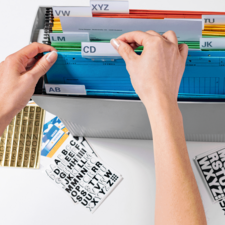 Hände sortieren farbige Ordner in einer Aufbewahrungsbox mit beschrifteten Registerkarten. Daneben liegen Etiketten und Aufkleber.