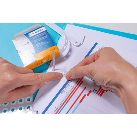 Eine Person befestigt eine transparente Kunststoffhülle an einem Dokument mit Diagrammen und Grafiken, während eine Verpackung im Hintergrund sichtbar ist.