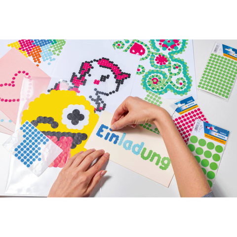 Hände gestalten Einladungen mit bunten Klebepunkten auf verschiedenen Papierblättern, darunter ein Monster und ein Herz.