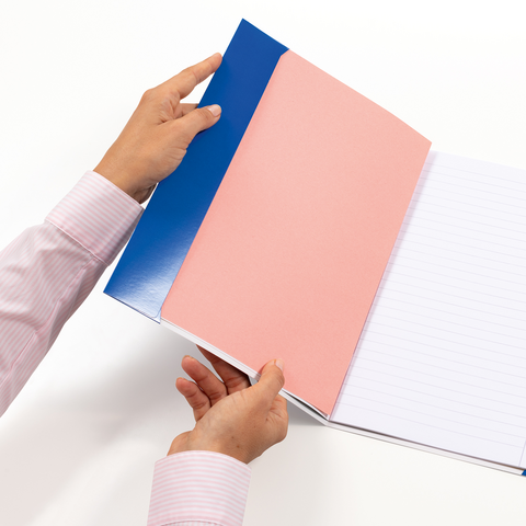 Zwei Hände halten ein geöffnetes Notizbuch mit einem blauen und einem rosa Einband. Die Seiten sind leer und liniiert.