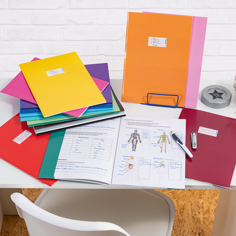Bunte Ordner und Dokumente auf einem Schreibtisch, daneben ein Glas mit Süßigkeiten und Bürobedarf wie einen Hefter und einen Stift.