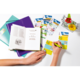 Eine Hand schreibt in ein Notizbuch, während mehrere bunte Karten und ein Blumenarrangement auf dem Tisch liegen.