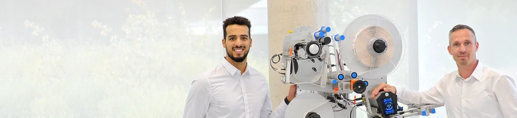 Zwei Männer in weißen Hemden stehen neben einer technischen Maschine mit verschiedenen Komponenten und Bedienelementen.