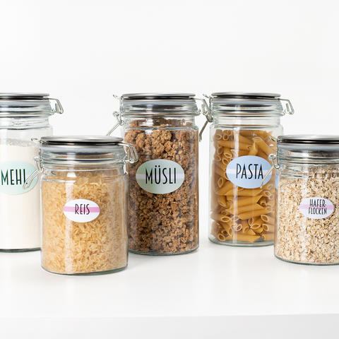 Fünf Glasbehälter mit Etiketten für Mehl, Reis, Müsli, Pasta und Haferflocken, auf einem weißen Tisch angeordnet.