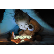 Ein Kind sitzt im Bett, umgeben von einer Decke, und hält ein leuchtendes Objekt in den Händen, während es einen Teddy umarmt.